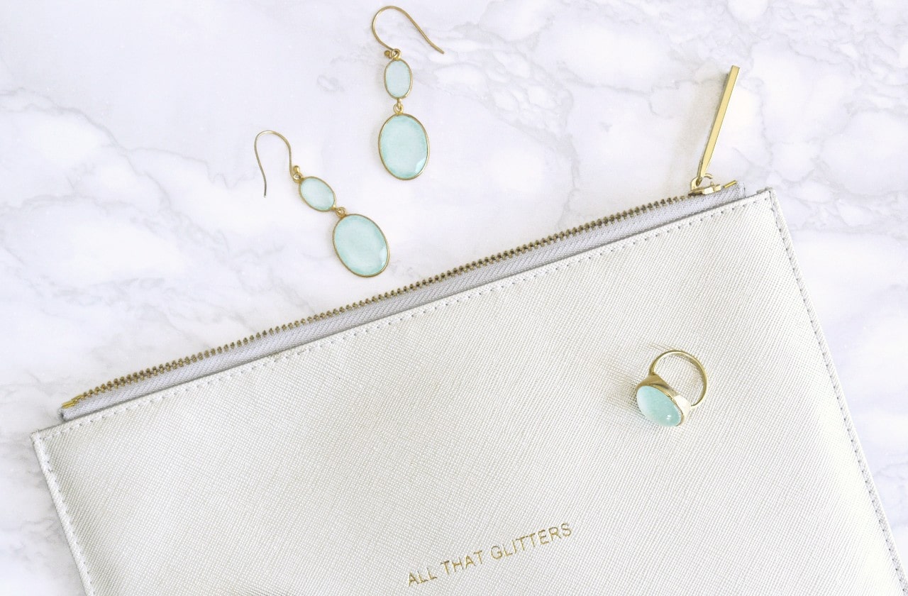 A pair of aquamarine and yellow gold earrings along with a matching fashion ring on top of a clutch on a white marble counter that says All That Glitters on it